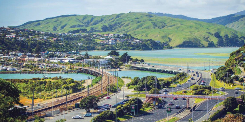 An image of a city and natural surroundings, with a railway, roads, bay, and green hills.